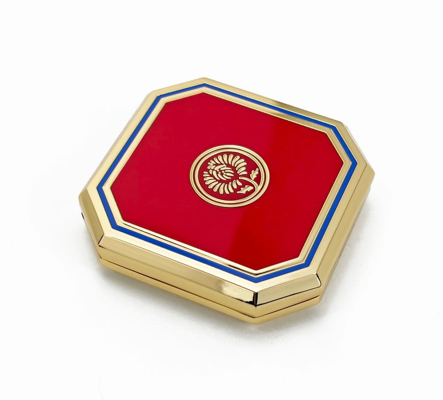 An octagonal compact case with a glossy red enamel center, bordered by a blue stripe and a polished gold-tone frame, featuring a gold emblem in the center.